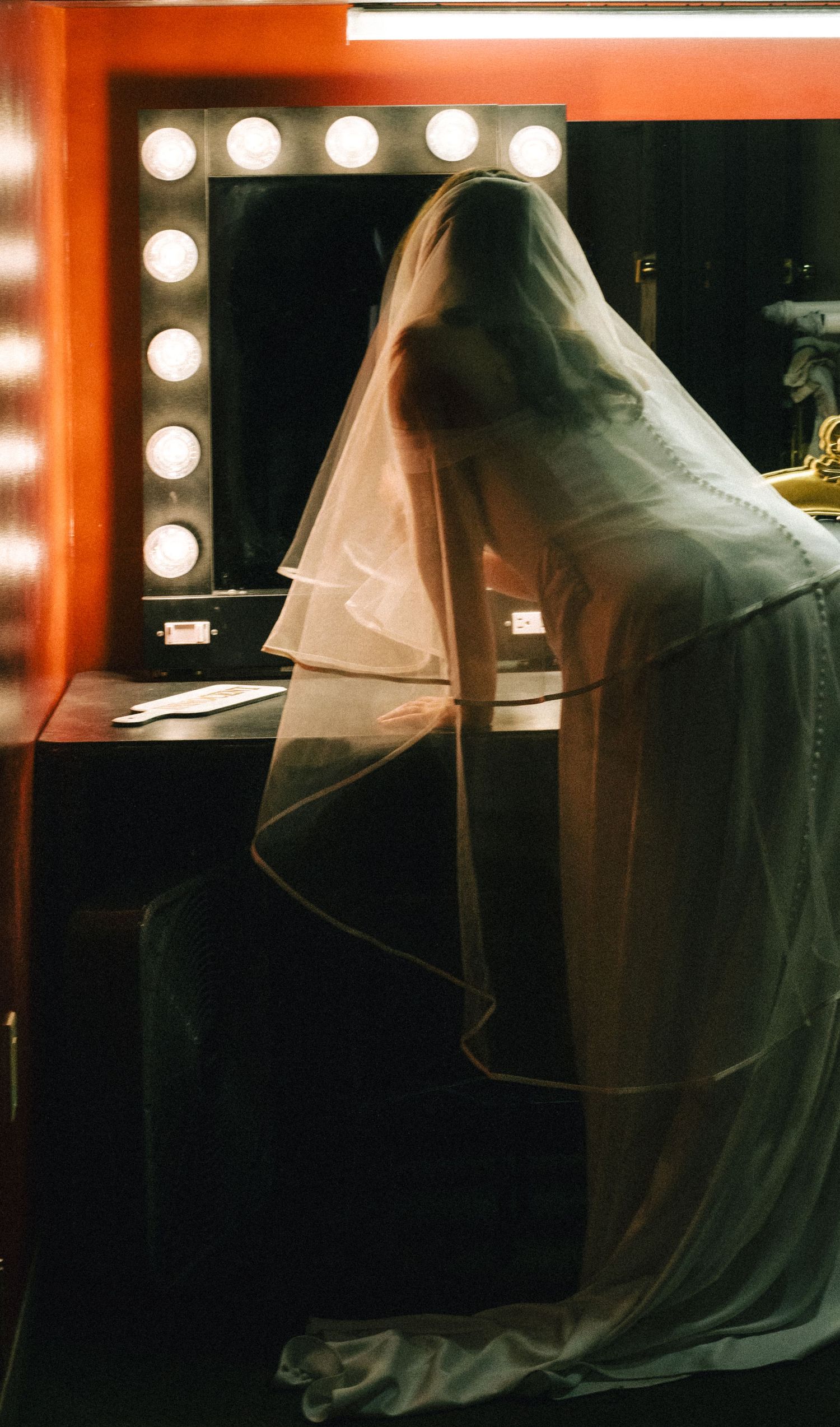 Bride in wedding veil sitting at vanity mirror with Hollywood lights.