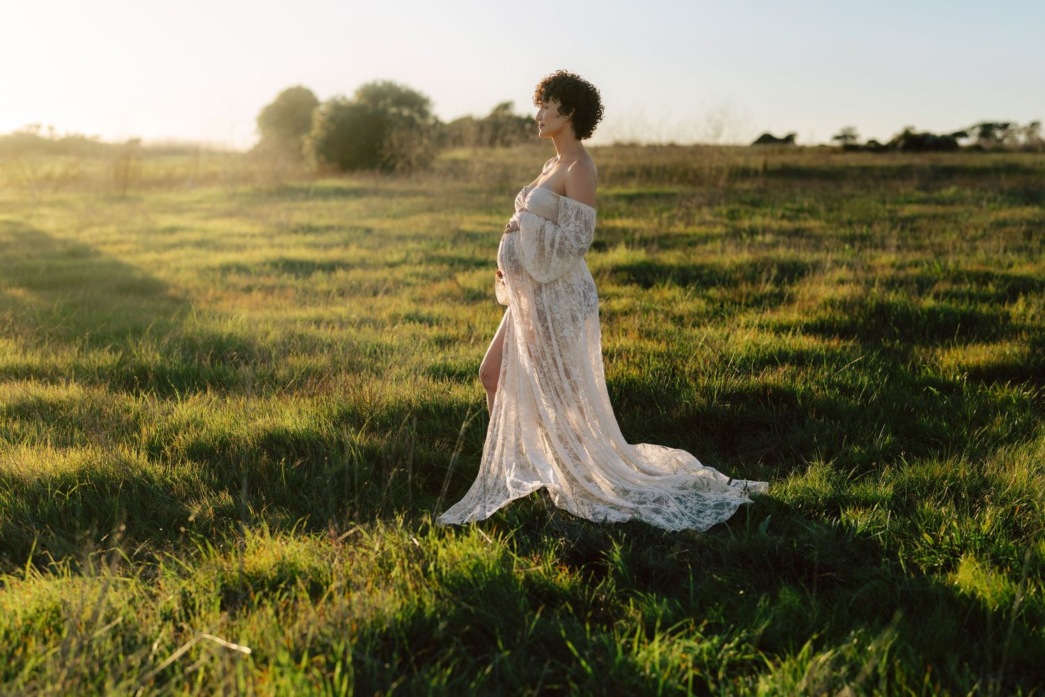 Pregnant woman in flowing white lace dress standing in sunlit green field.