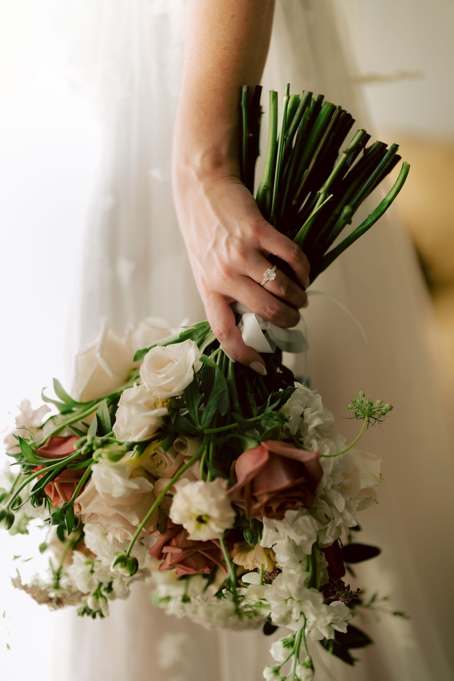 Bride holding a lush bridal bouquet of white and blush roses with a diamond engagement ring visible.