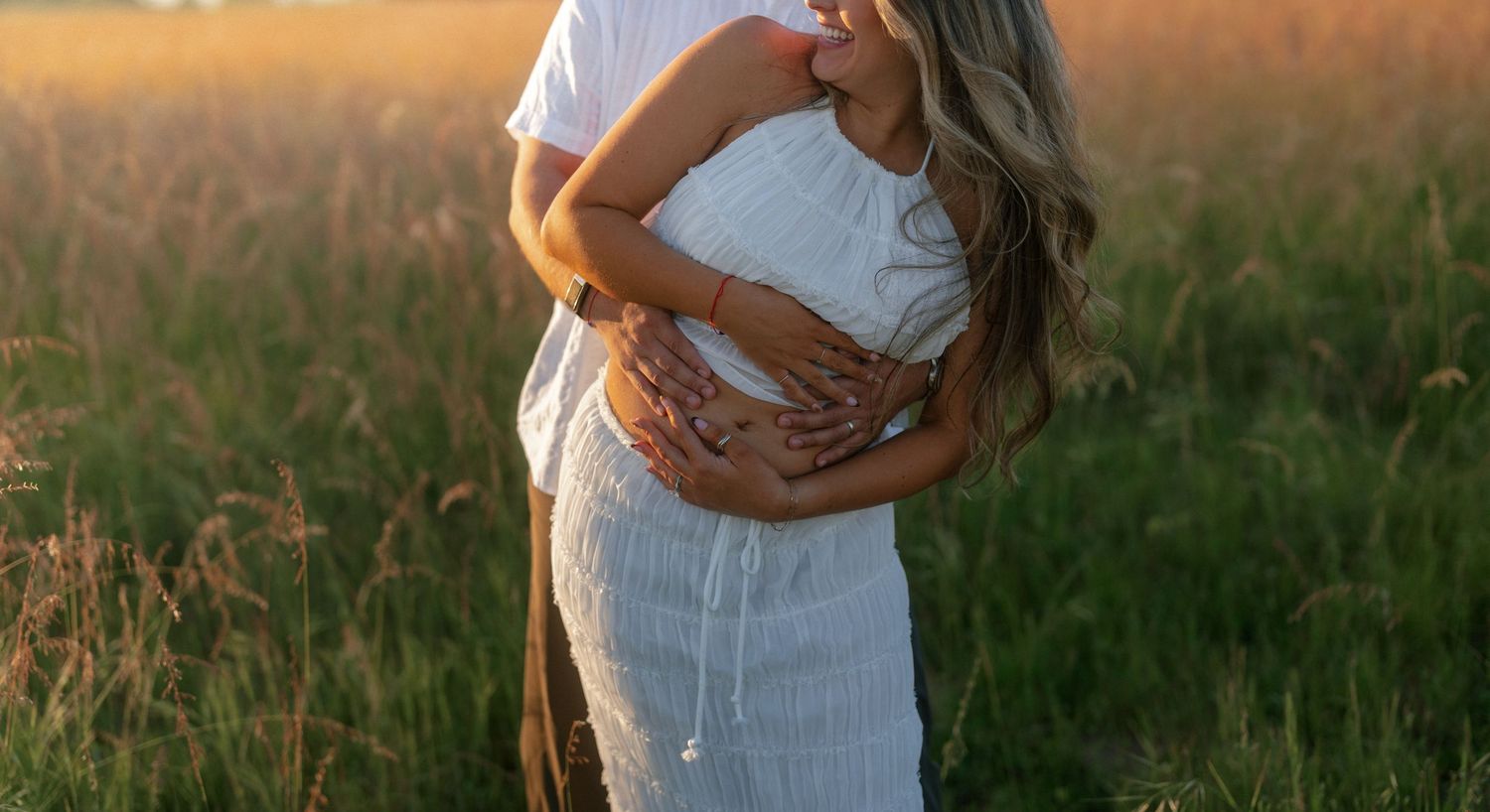 Pregnant couple embracing in golden sunlit field, woman in white dress with hands cradling baby bump.