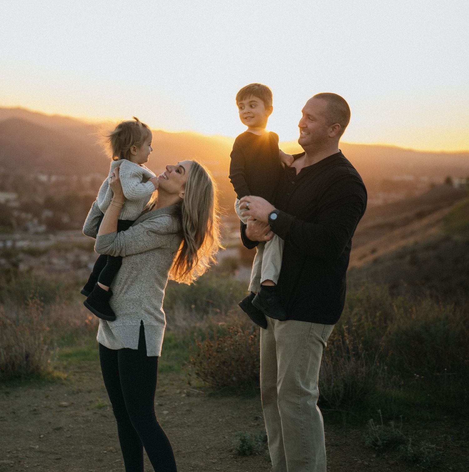 Family of four at golden hour sunset, parents lifting two children on a scenic hillside.