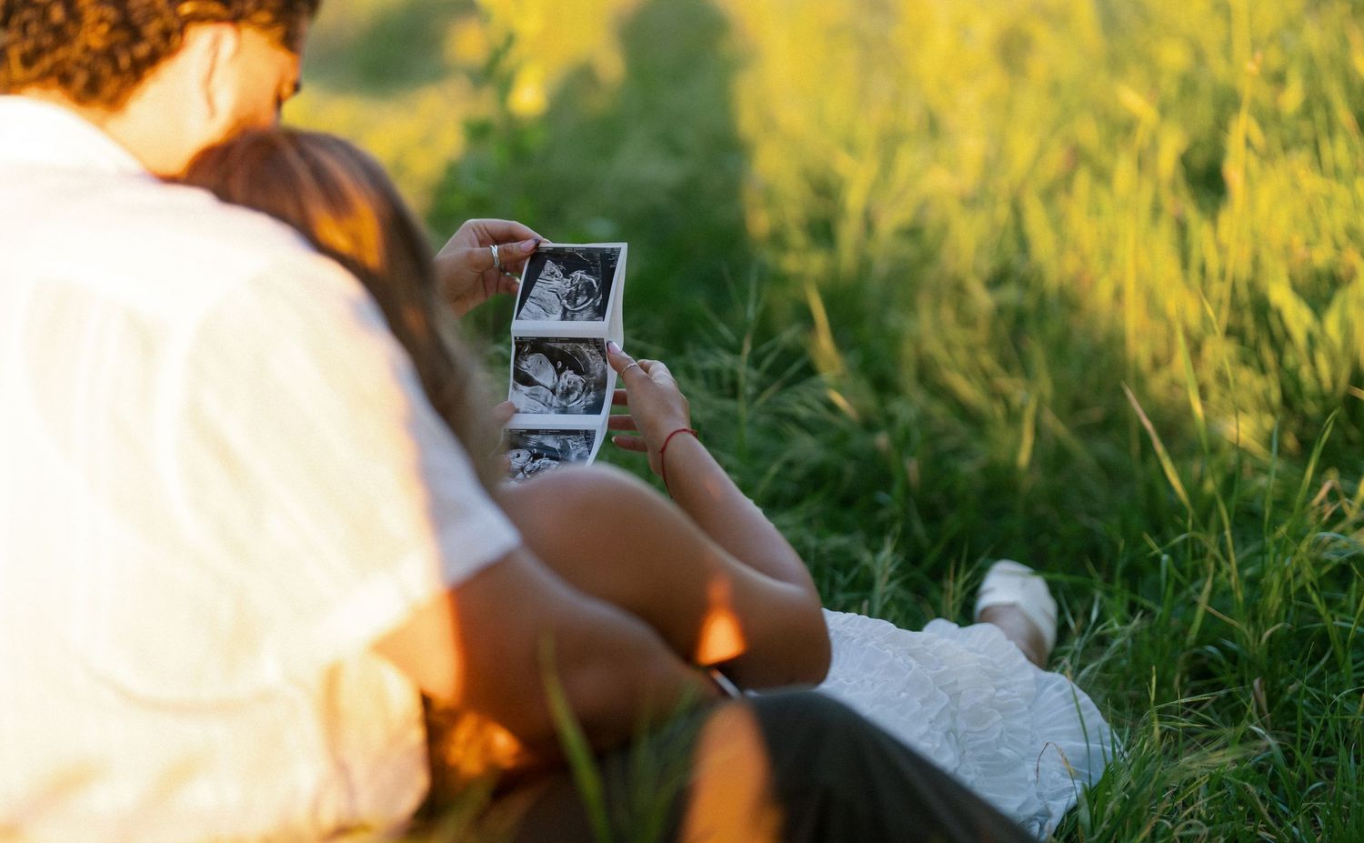 Couple sitting in green grass field holding ultrasound photos, announcing pregnancy in golden sunset light.