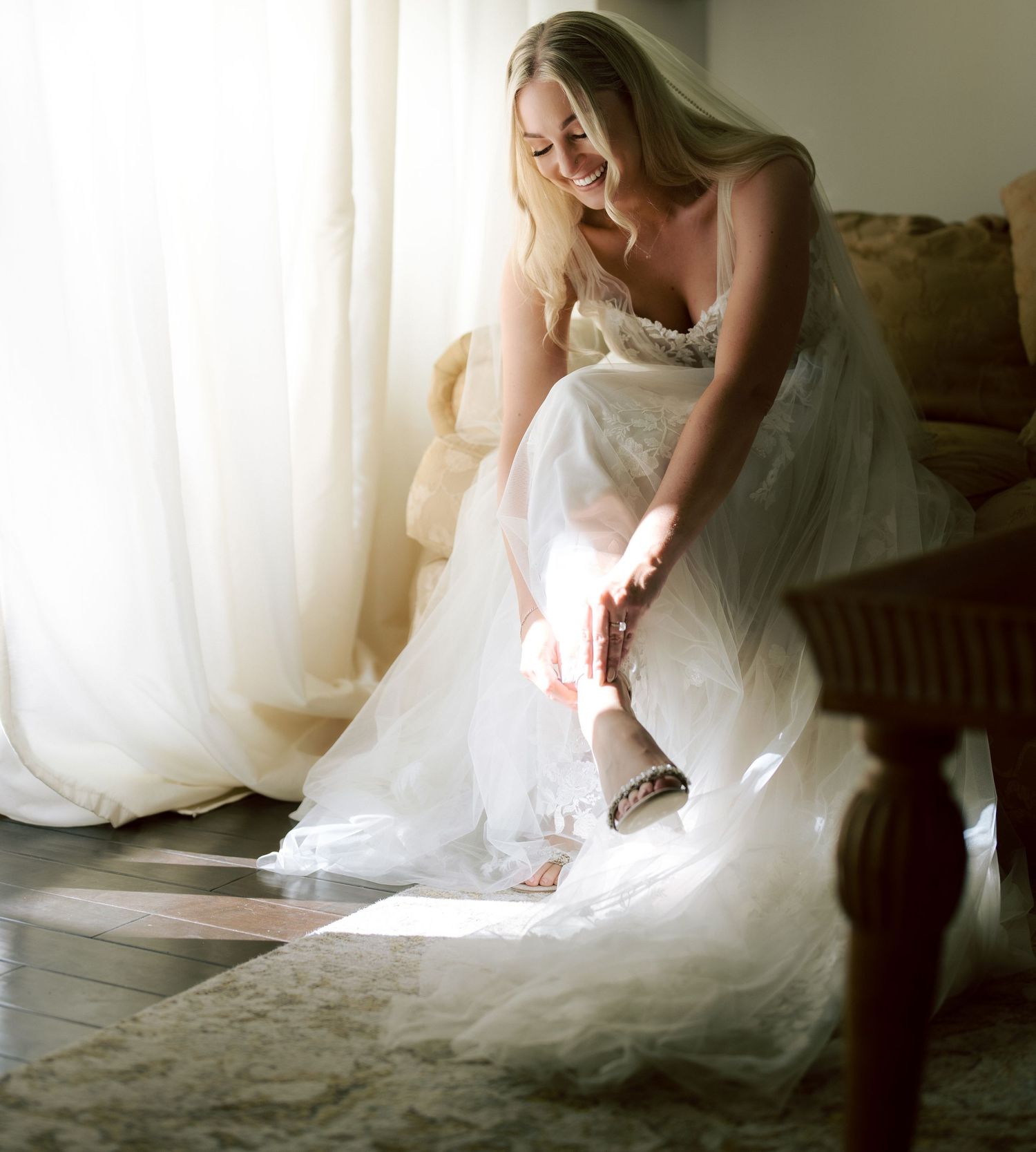 Blonde bride in a white lace gown putting on her shoes in a sunlit room before the wedding.