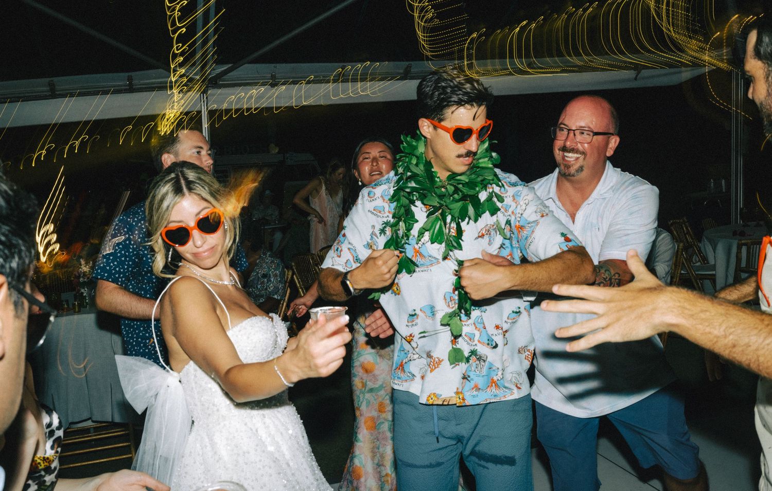 Bride and groom in Hawaiian attire dancing joyfully with guests at a tropical outdoor wedding reception.