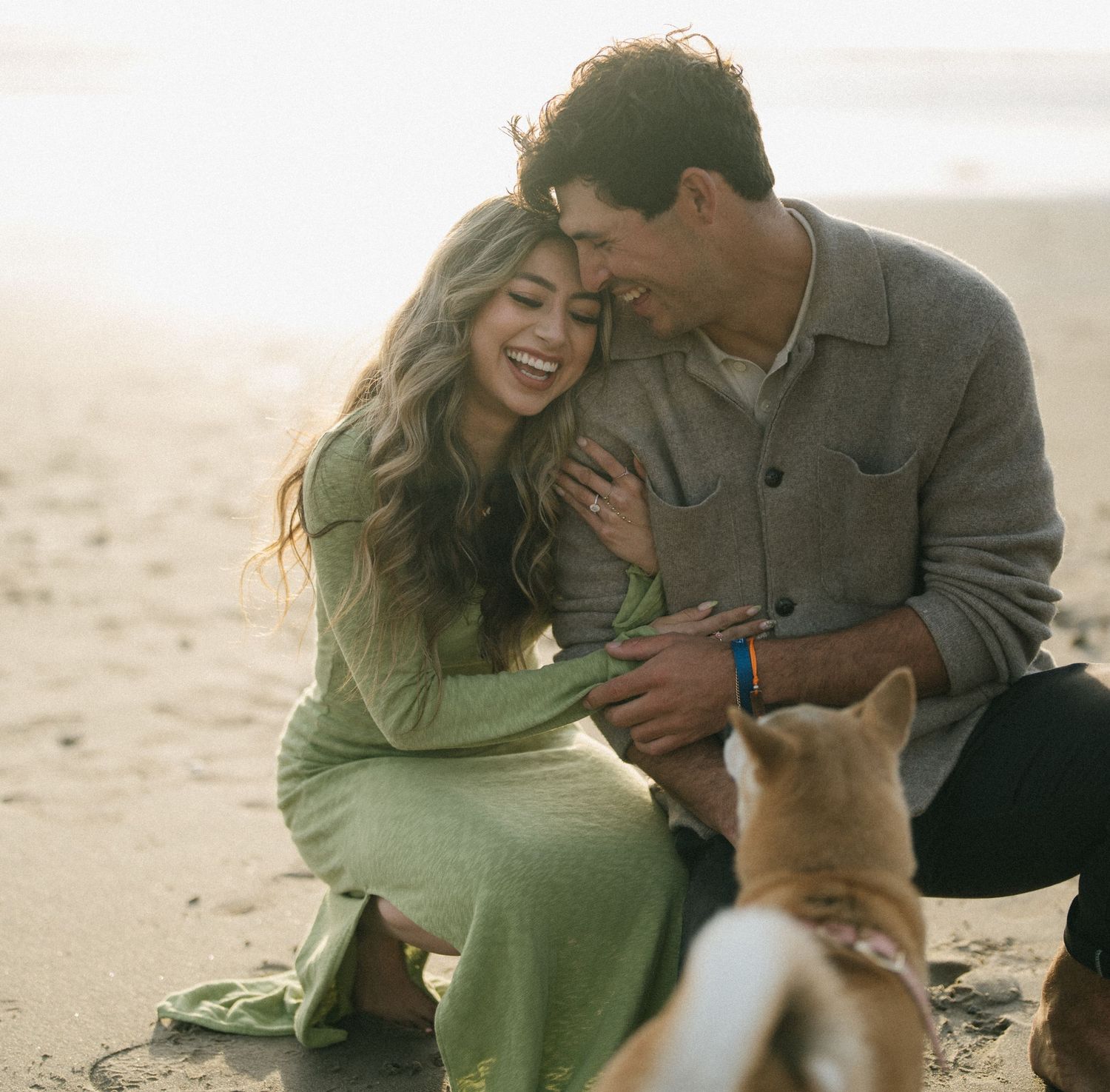 Laughing couple crouching on a sandy beach petting a Corgi dog in warm sunset light.