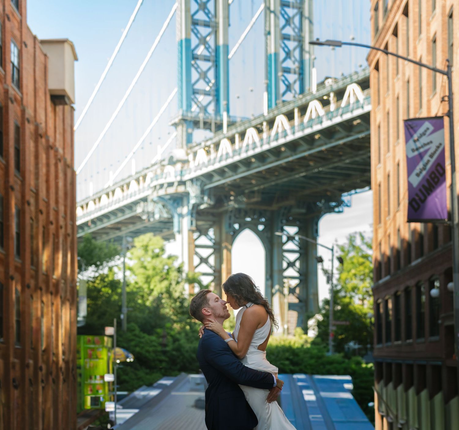 Couple sharing a romantic kiss in DUMBO Brooklyn with the Manhattan Bridge framed between brick buildings.