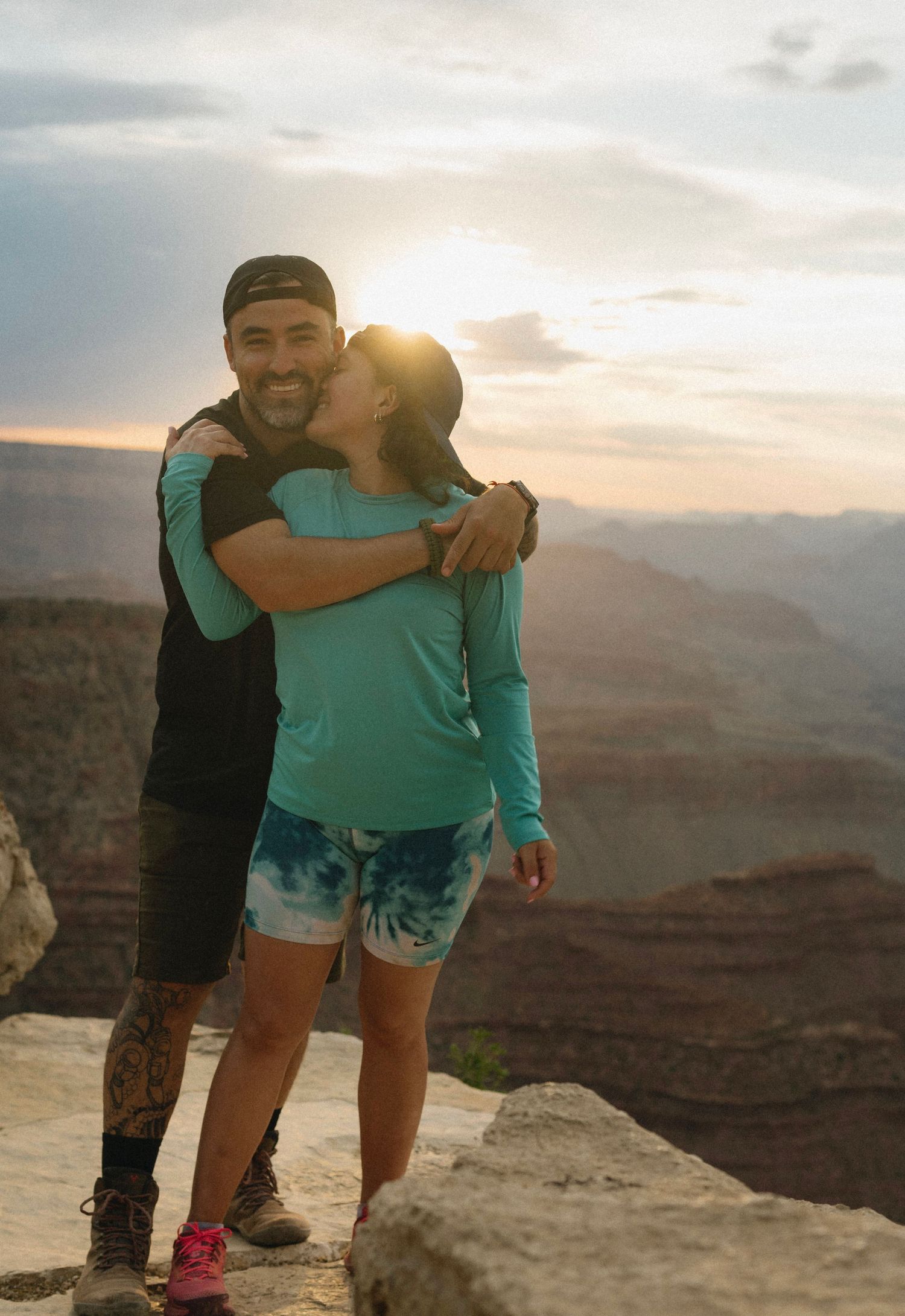 Couple hugging and smiling at sunset over Grand Canyon with dramatic sky backdrop.