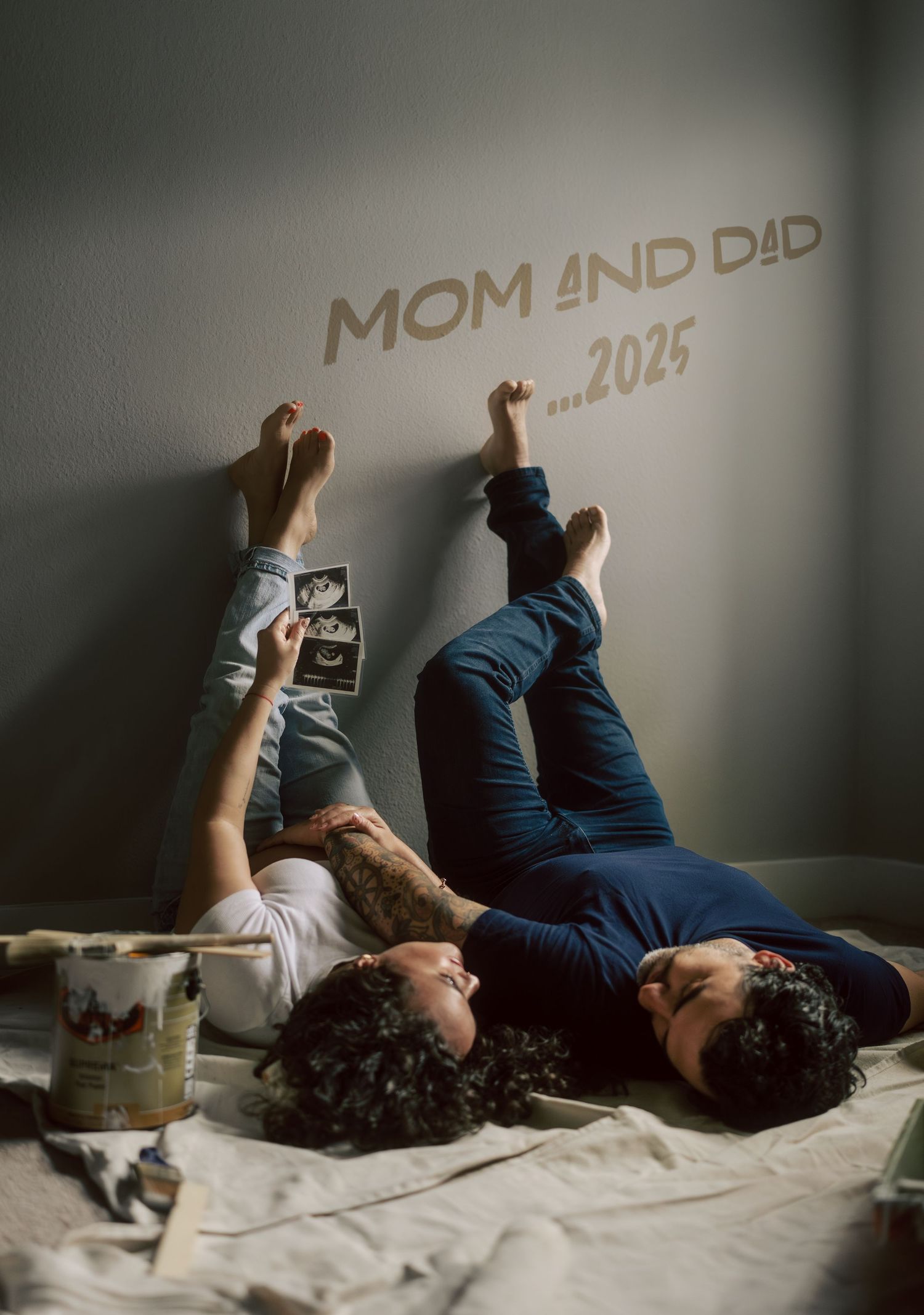 Couple lying on floor holding ultrasound photos beneath Mom and Dad 2025 wall art.