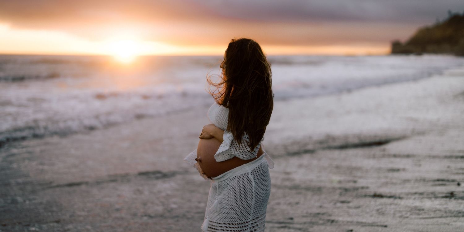Pregnant woman in white dress standing on beach at dramatic sunset.