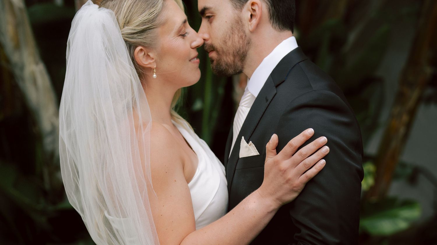 Bride and groom share an intimate romantic moment, nearly kissing, surrounded by lush greenery.