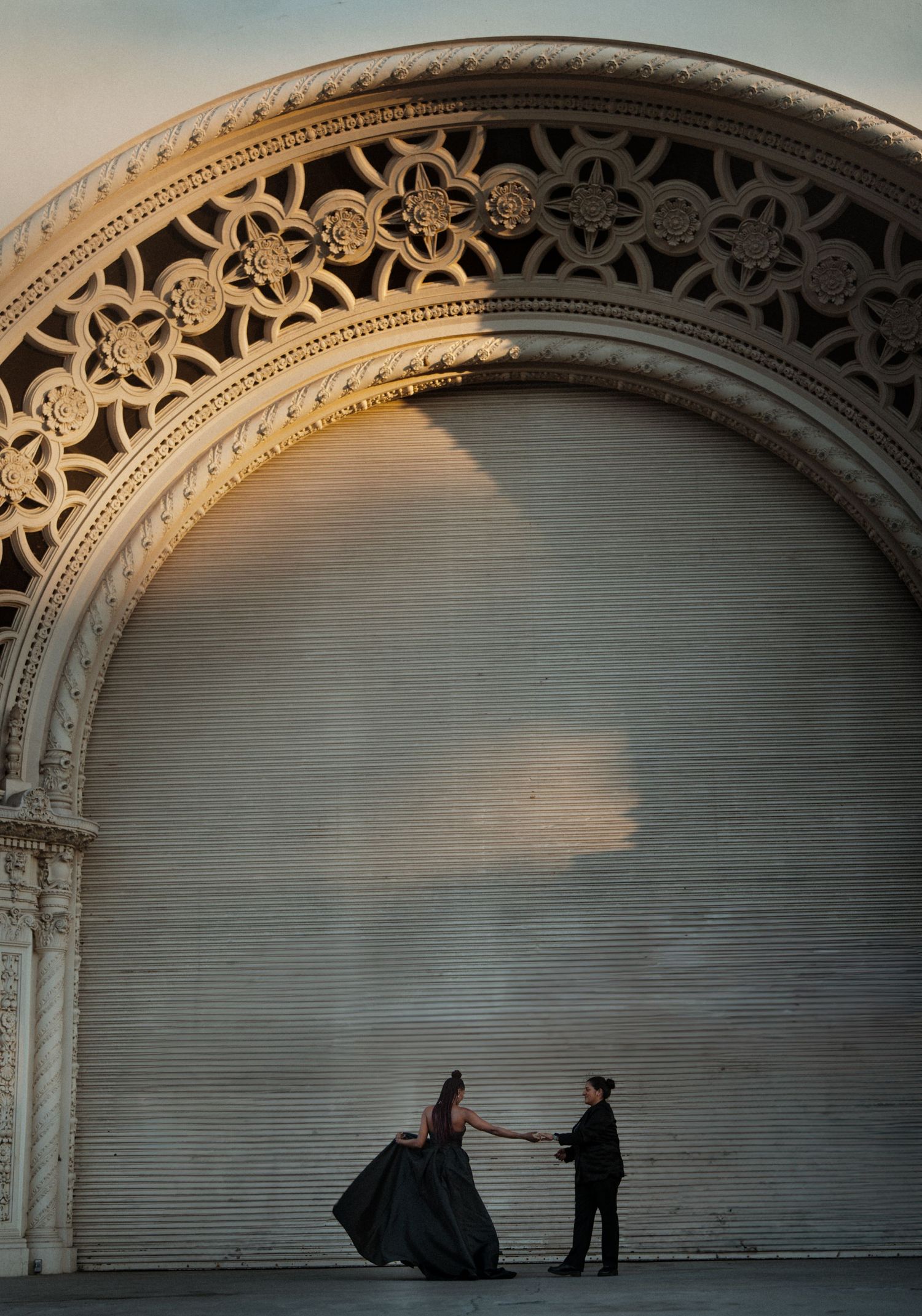 Couple dancing in black formal wear beneath an ornate decorative arch at golden hour.