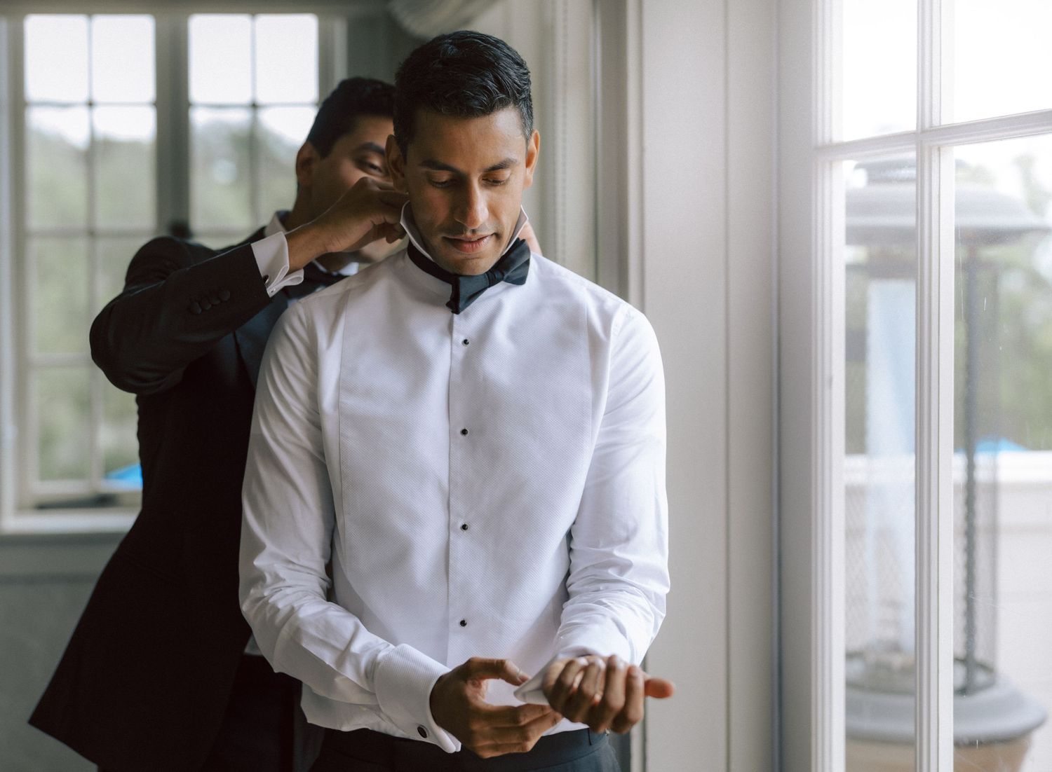 Groom in white shirt gets bow tie adjusted by groomsman near bright window before wedding.
