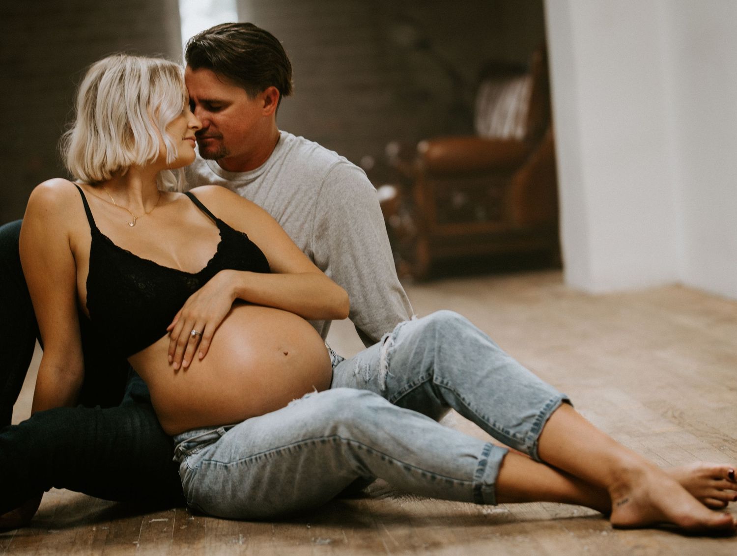 Expecting couple sitting on wooden floor, man tenderly kissing pregnant woman while holding her baby bump.