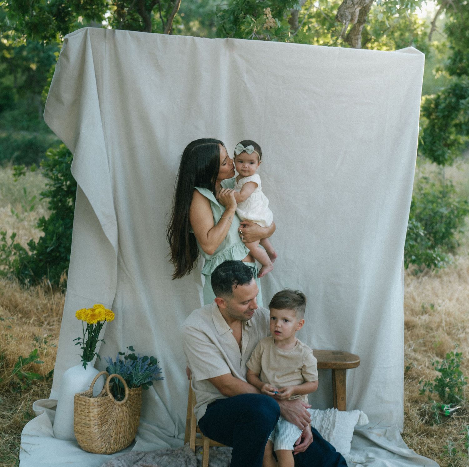 Mother kissing baby while father sits with son outdoors against a white linen backdrop.