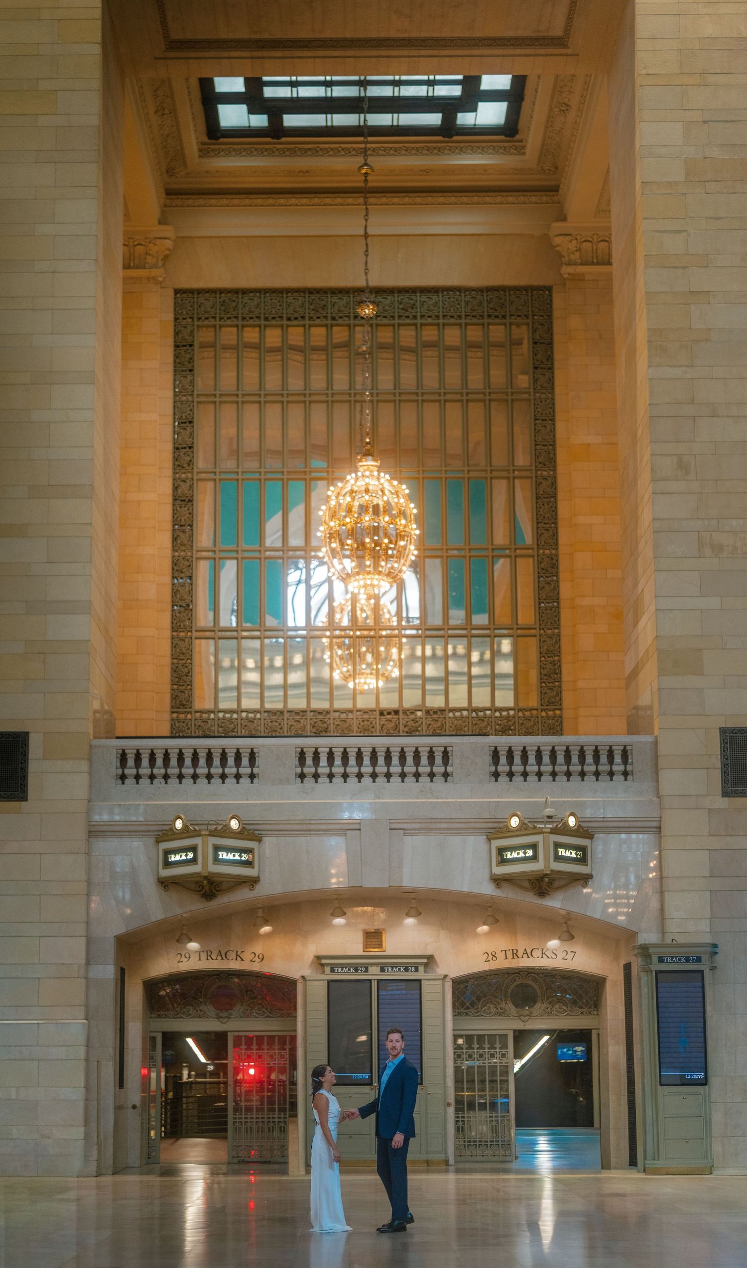 Bride and groom standing beneath a grand chandelier in the elegant main concourse of Grand Central Terminal, New York.