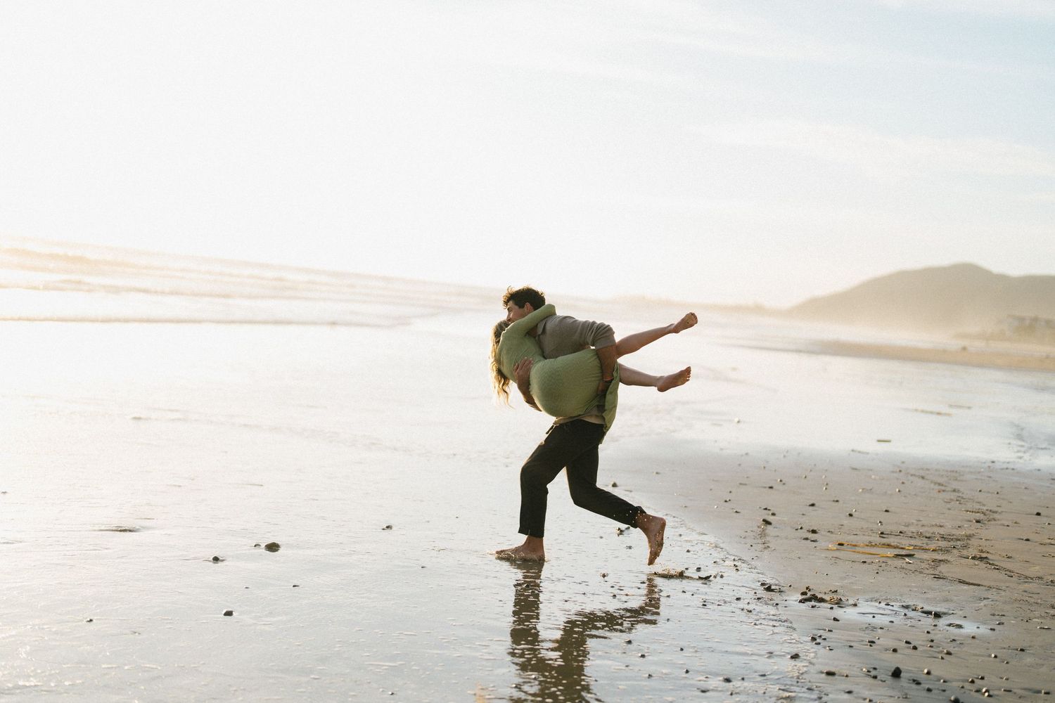 Man giving woman a piggyback ride while running playfully along a sunny reflective beach.