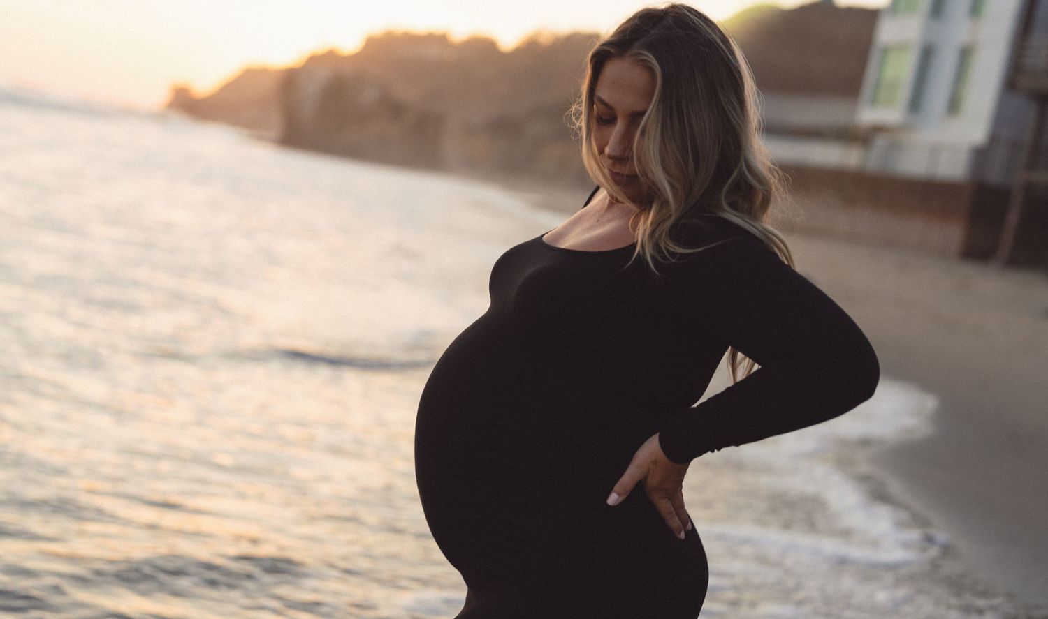 Pregnant woman in black dress holding belly on beach at sunset.