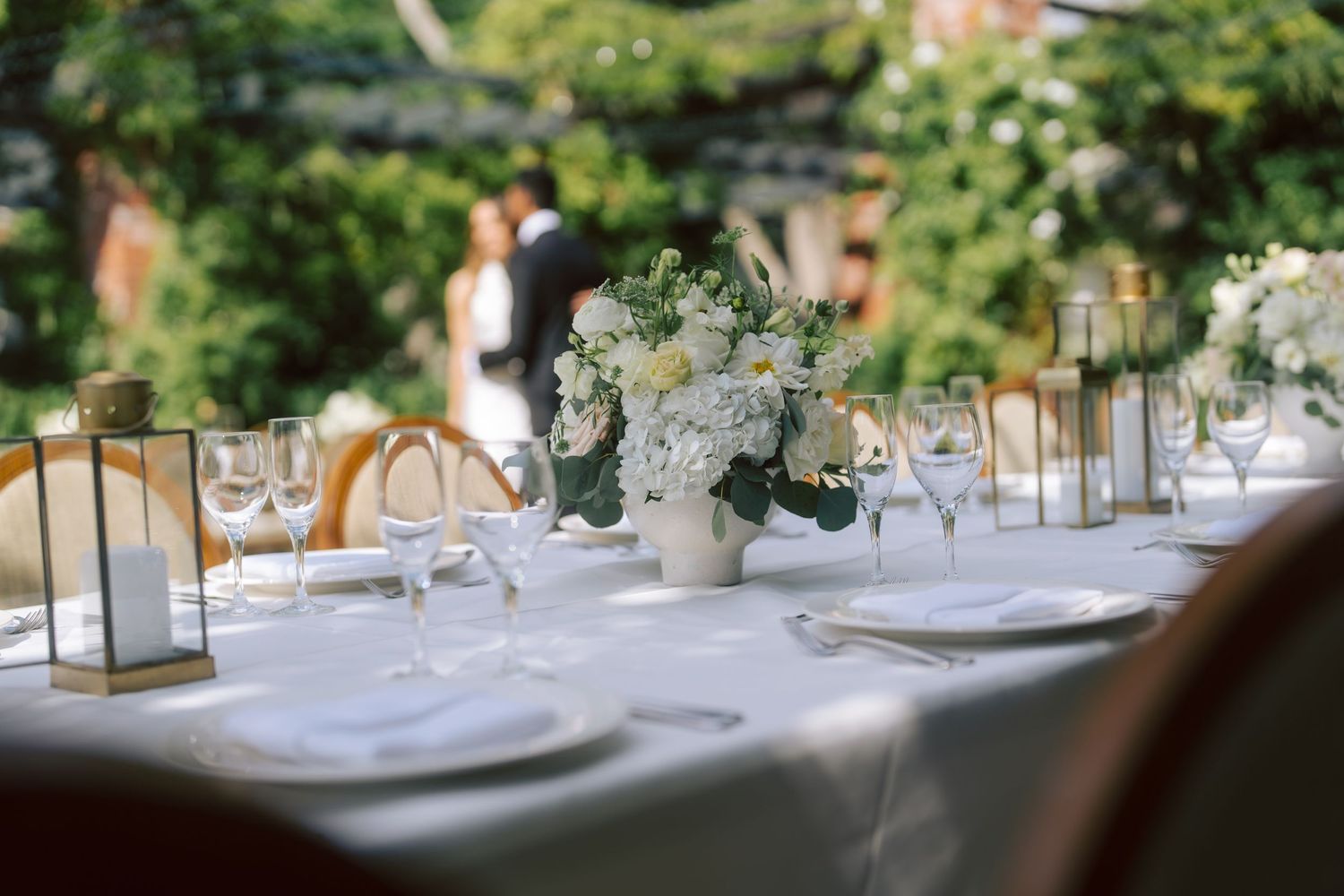 Elegant outdoor wedding reception table with white floral centerpiece, candles, and glassware set among lush greenery.