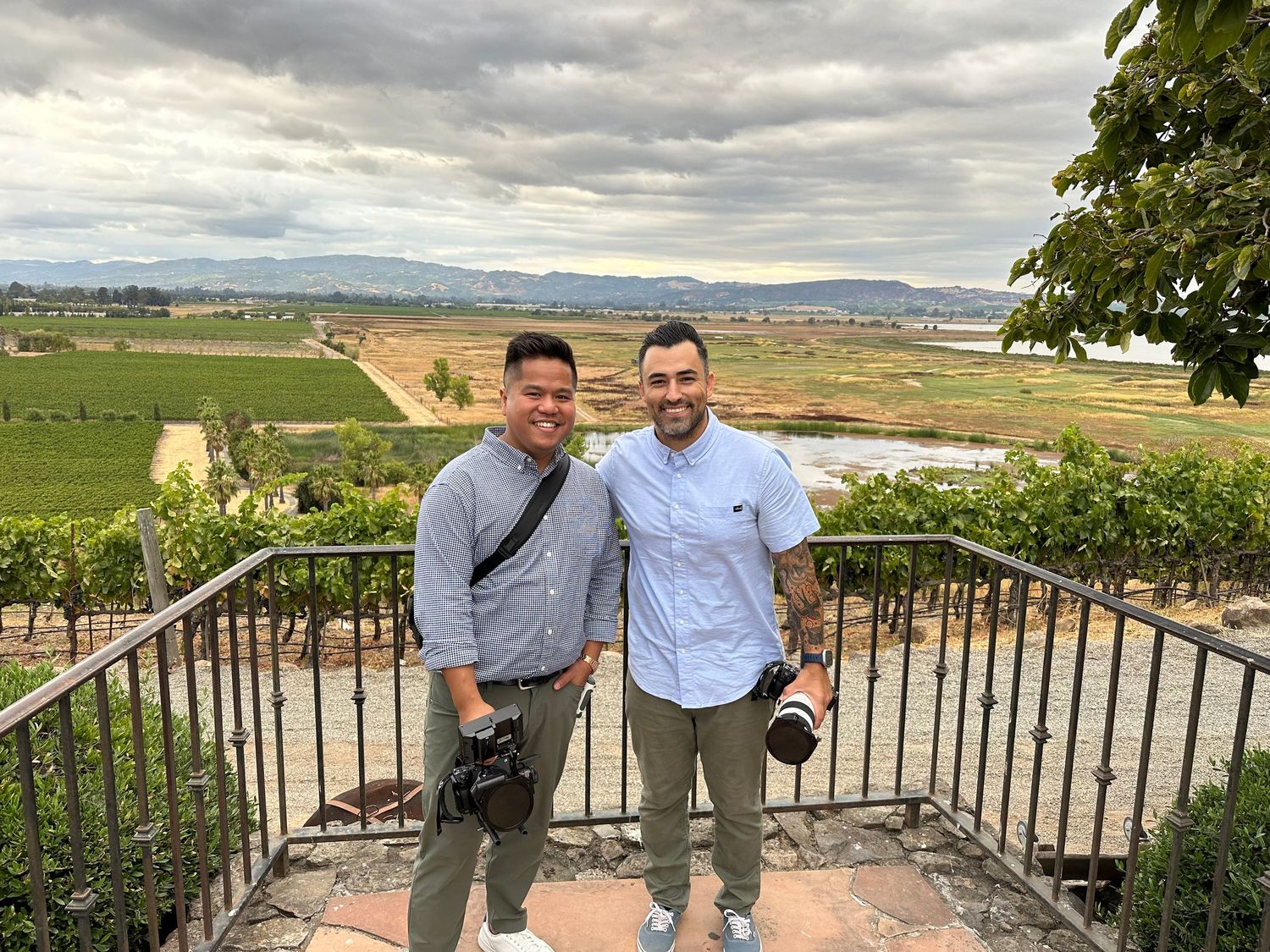 Two men smiling at a scenic vineyard overlook with lush green fields and cloudy skies in the background.