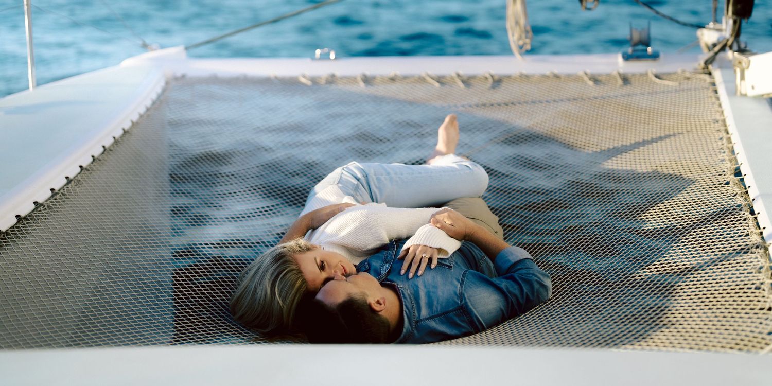 Romantic couple lying together on the net of a catamaran sailboat over turquoise ocean waters.