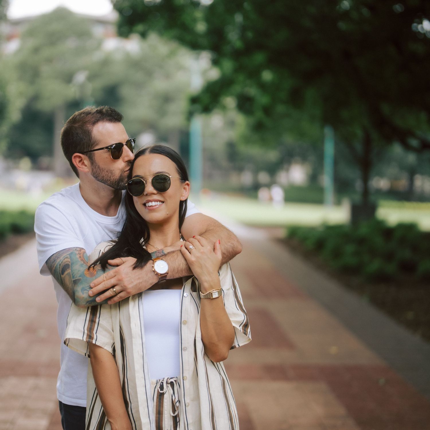 Couple embracing in a sunny park, man kissing woman's cheek, both wearing sunglasses and casual stylish outfits.