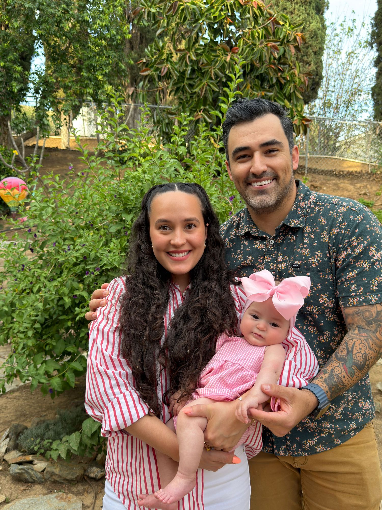 Happy family portrait with parents holding their baby girl outdoors in a garden setting.