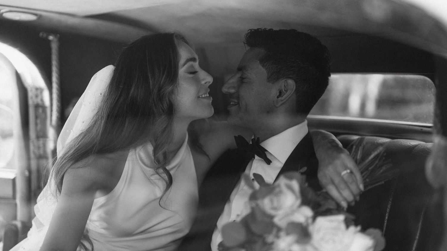 Black and white close-up of a smiling bride and groom sharing an intimate moment in a vintage car.