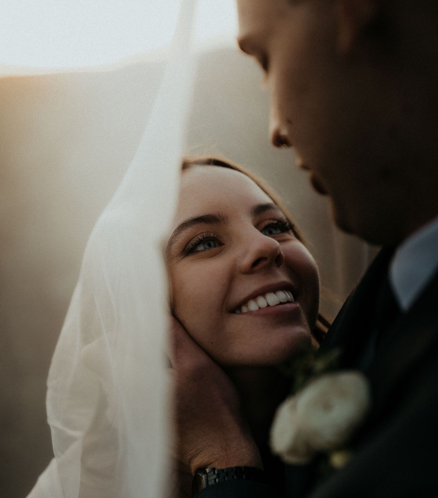 Radiant bride smiling under her veil at her groom in a golden sunlit wedding portrait.