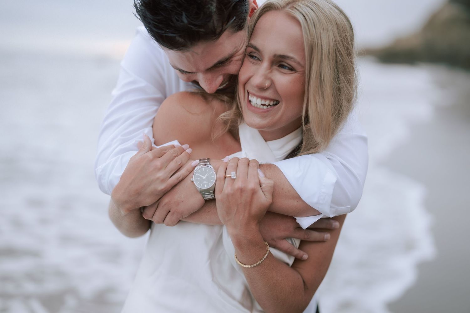 Joyful couple hugging tightly on beach, woman smiling and showing engagement ring on her finger.