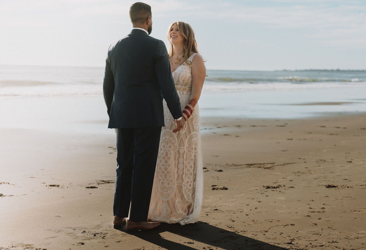 Bride and groom hold hands on a sandy beach in soft natural light after wedding.