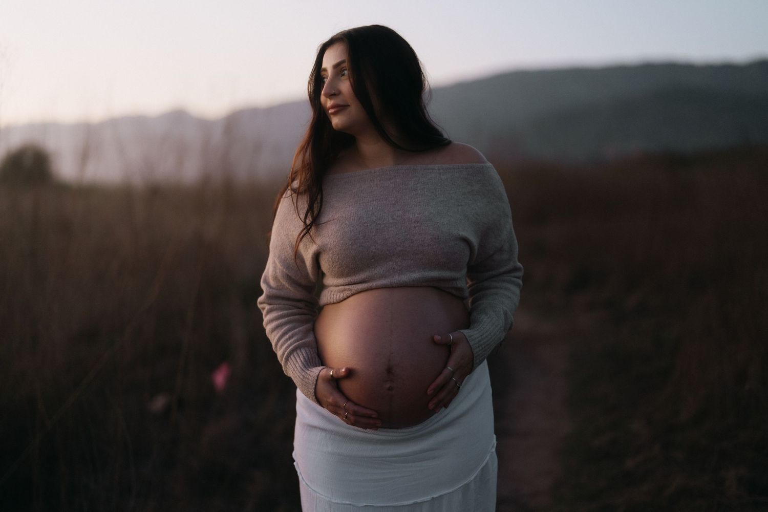 Pregnant woman cradling her baby bump in a moody dusk field with mountain backdrop.