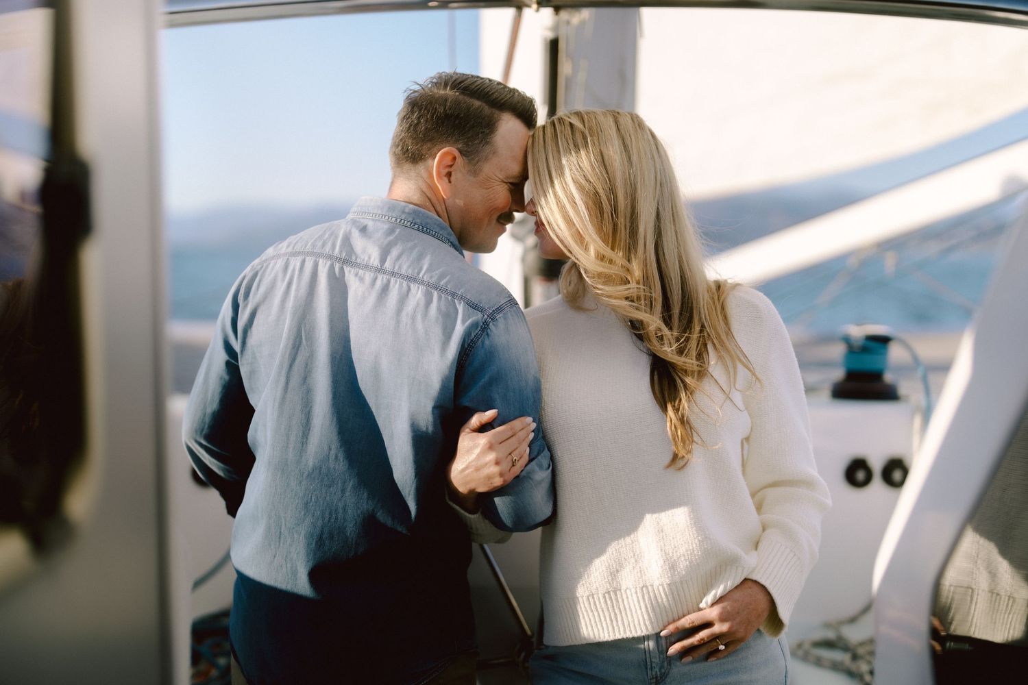 Couple sharing a tender nose-to-nose moment aboard a sailboat with scenic mountain water views behind them.