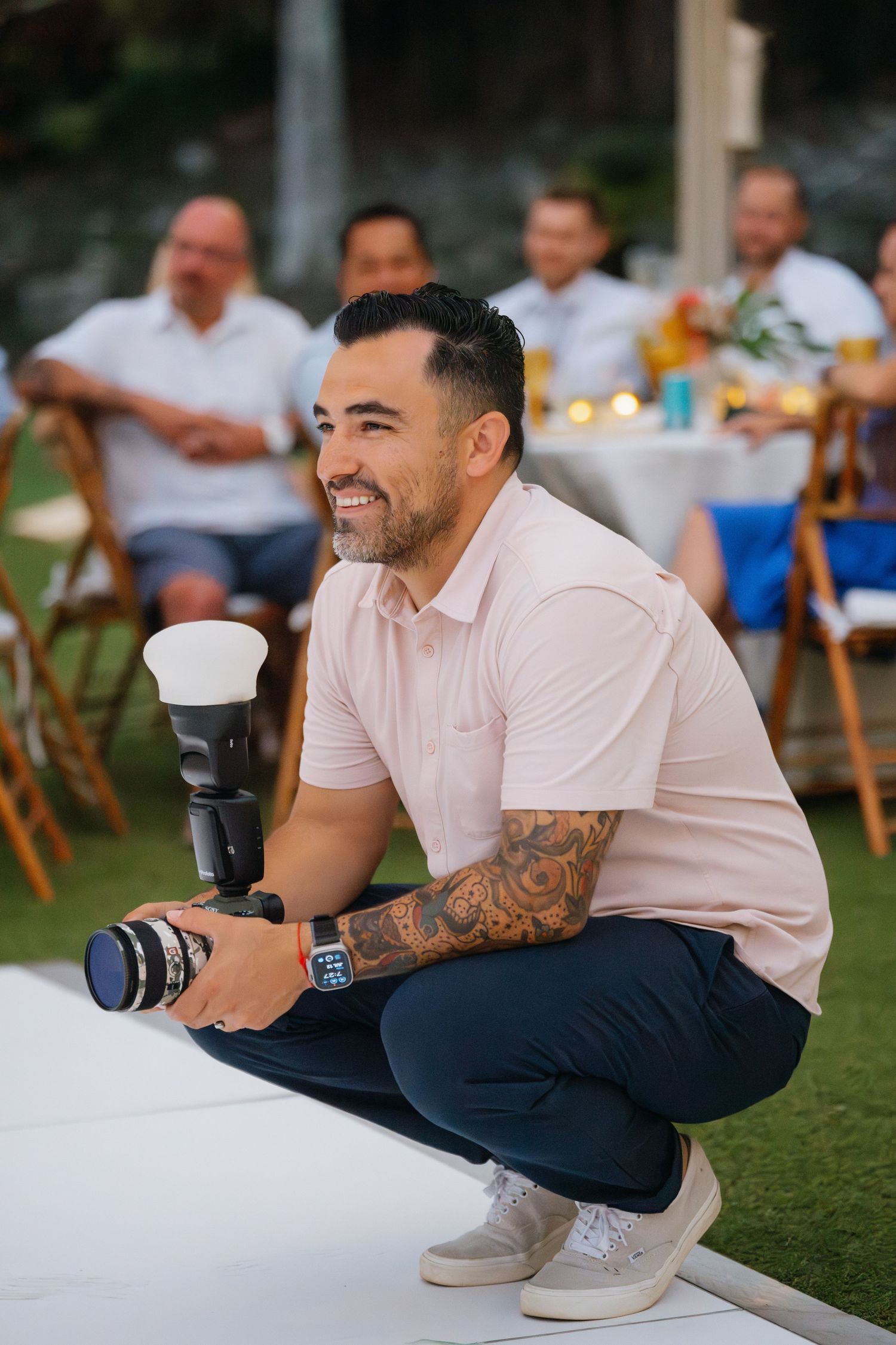 Tattooed man crouching with camera at outdoor wedding event smiling joyfully.