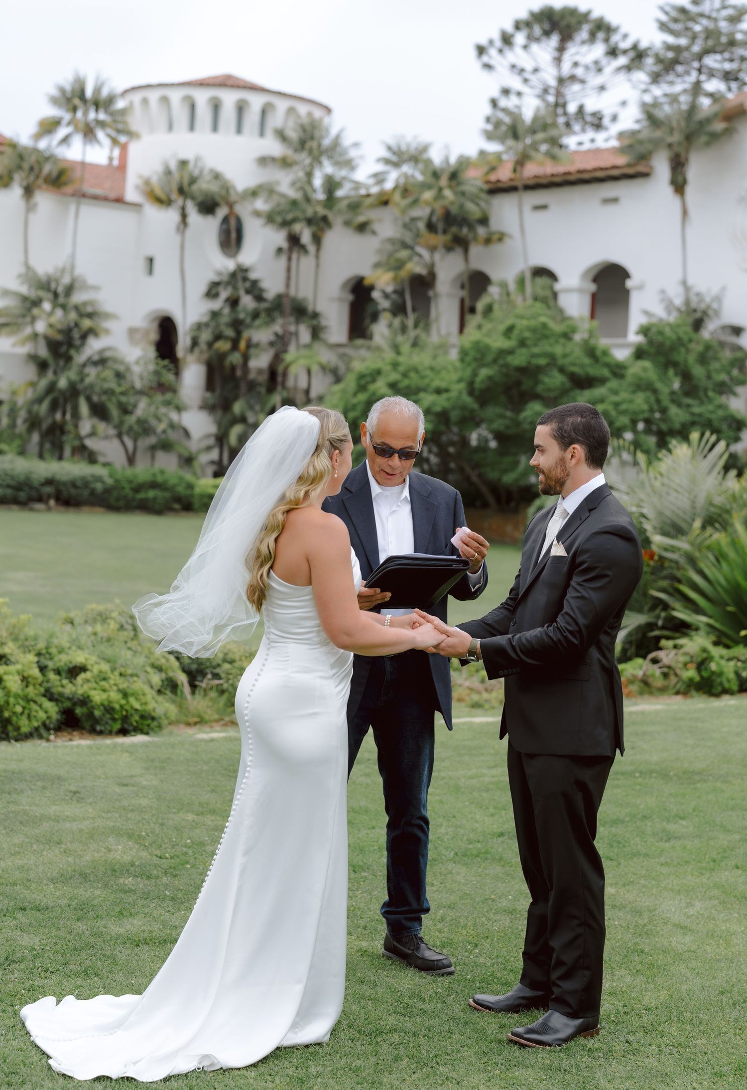 Bride and groom hold hands during outdoor wedding ceremony officiated before a Spanish colonial building.