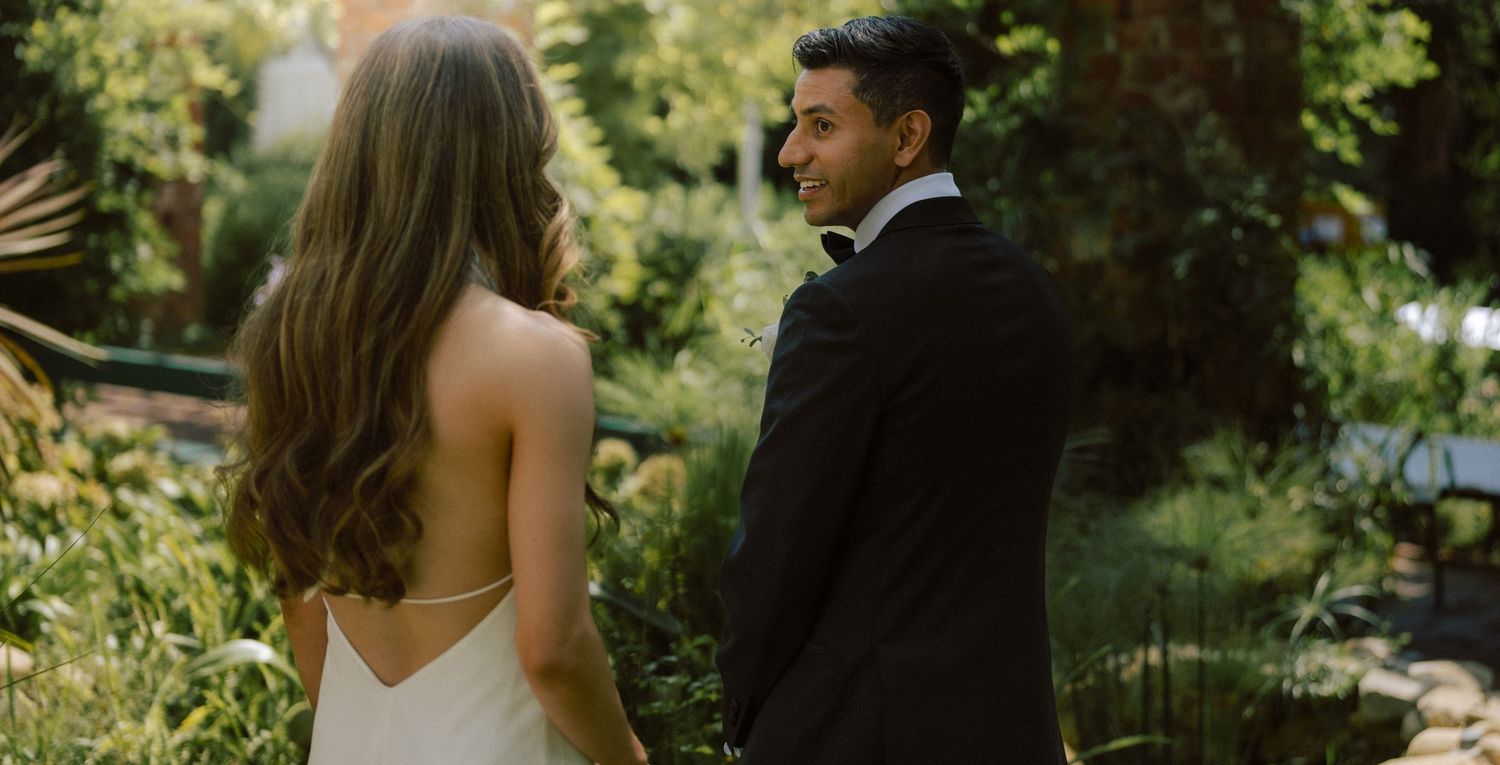 Groom in black tuxedo smiles at bride in white slip gown among lush garden greenery.