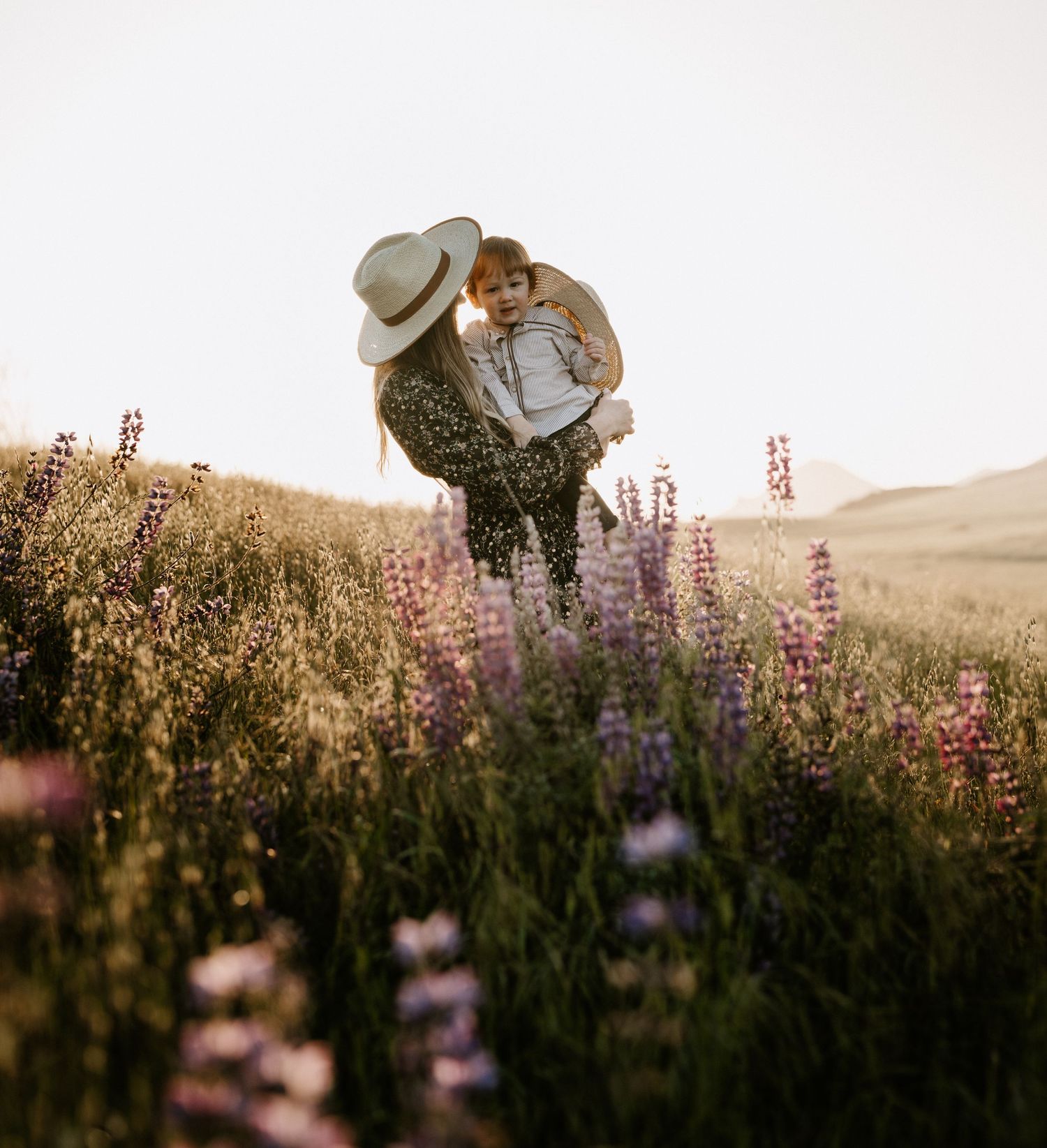 Mother in a straw hat holding her young child in a vibrant wildflower field at sunset.