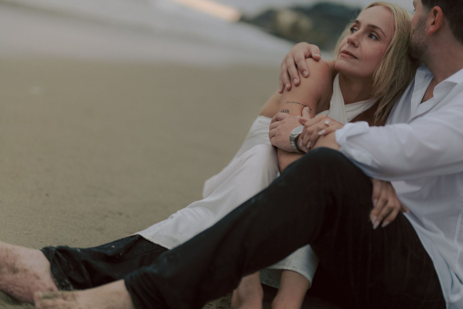 Couple sitting intimately on sandy beach at sunset, woman leaning back against man in white shirt.
