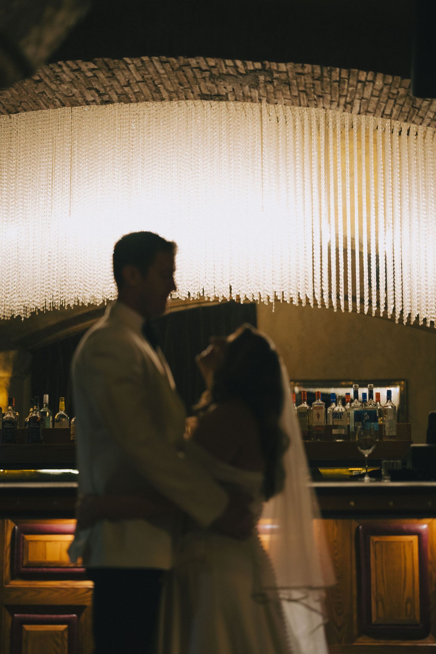 Bride and groom share romantic first dance silhouette in front of glowing chandelier at elegant wedding reception bar.