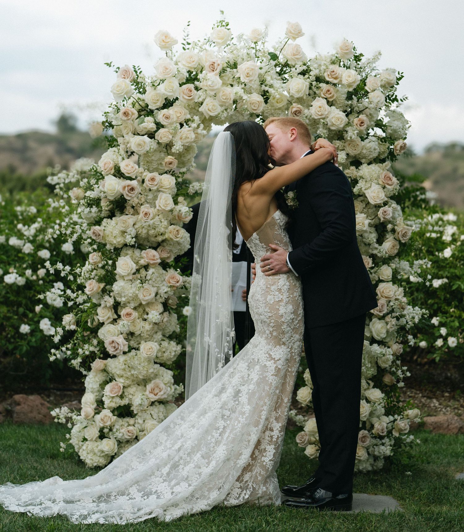 Couple shares first kiss at altar beneath stunning white floral arch at outdoor wedding.