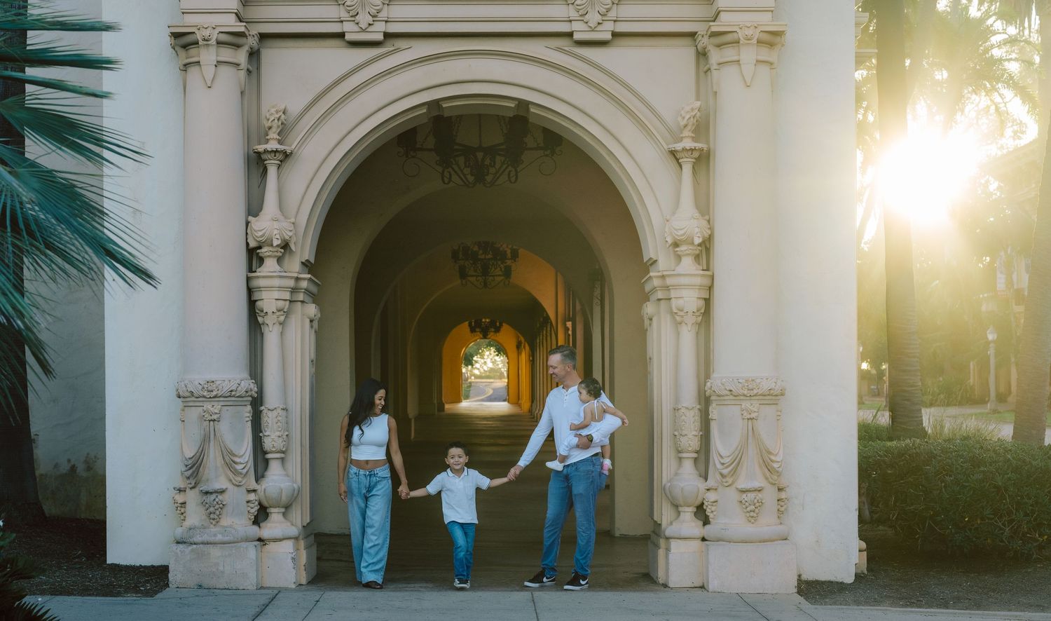 Family of four holding hands walking through ornate arched entrance in warm sunlight.
