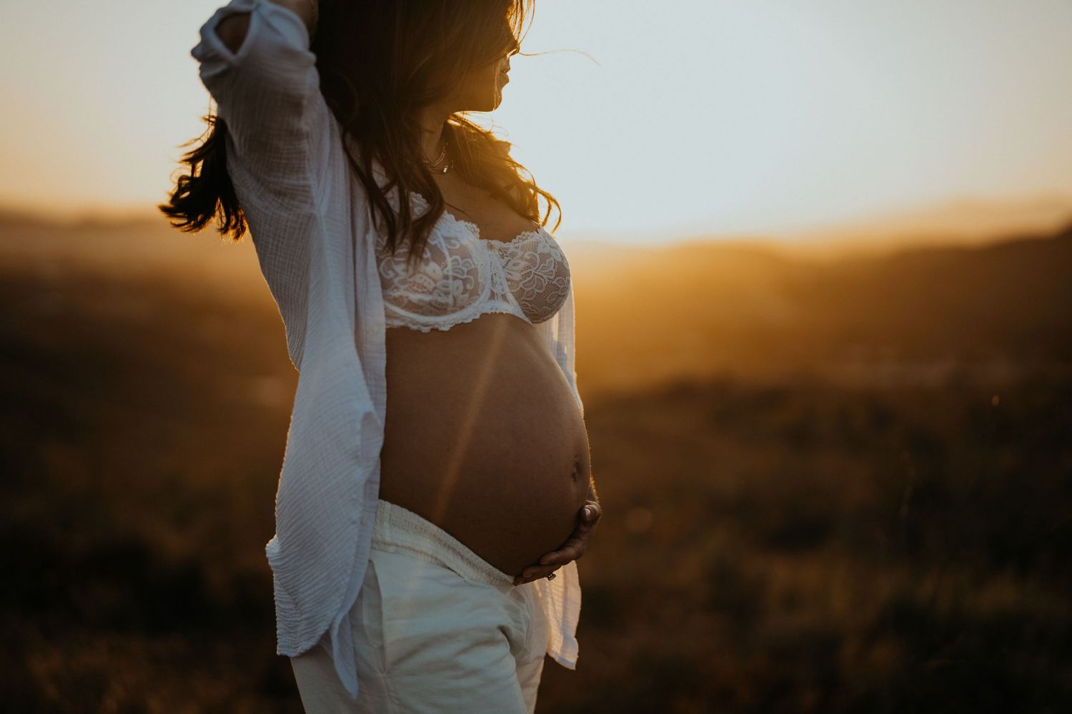 Pregnant woman in white outfit showing baby bump while standing in a glowing golden sunset field.