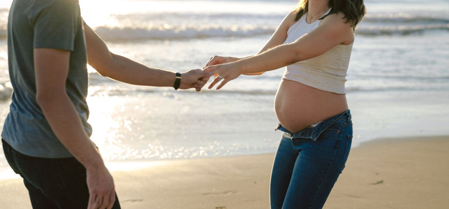 Pregnant couple holding hands on sunny beach, woman in white top and jeans.