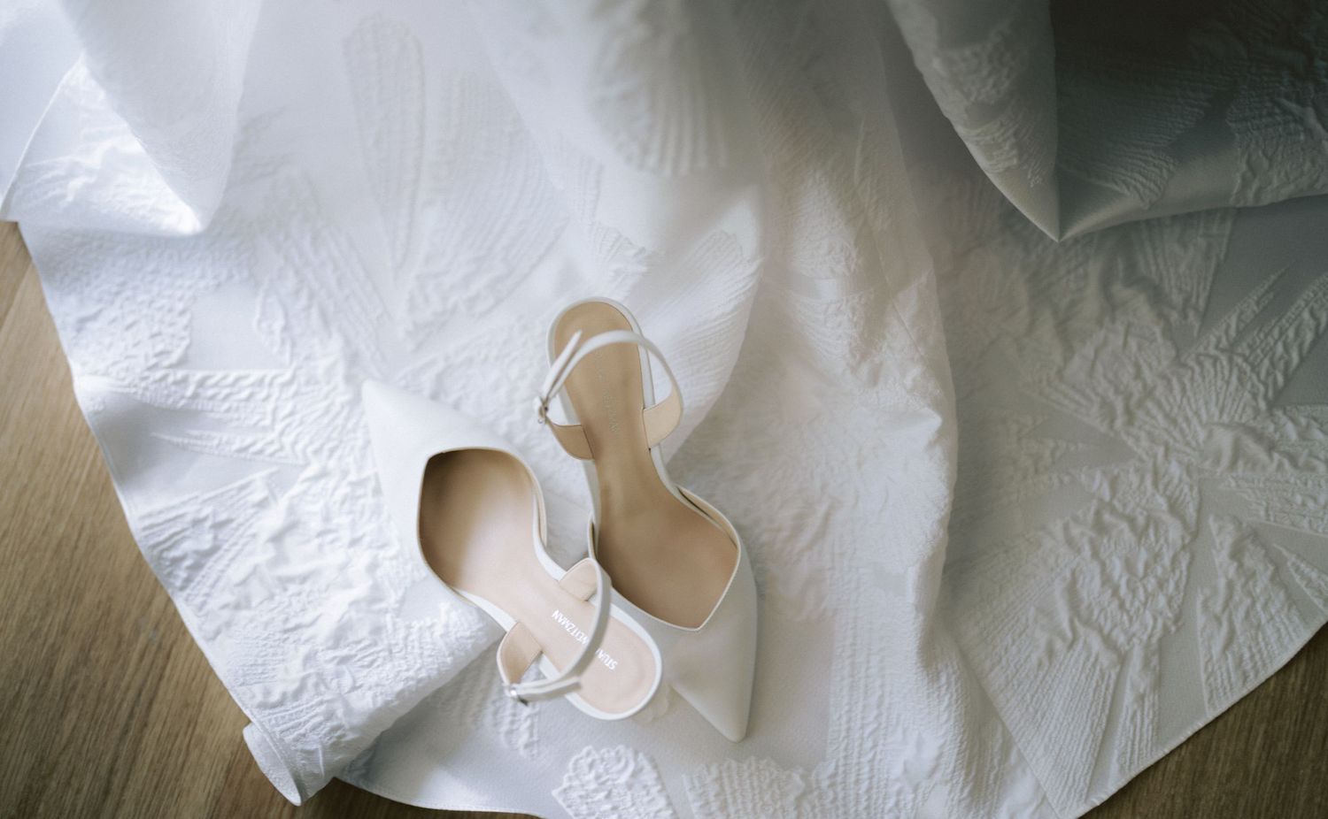 White strappy bridal heels resting on embroidered white wedding gown fabric on wooden floor.