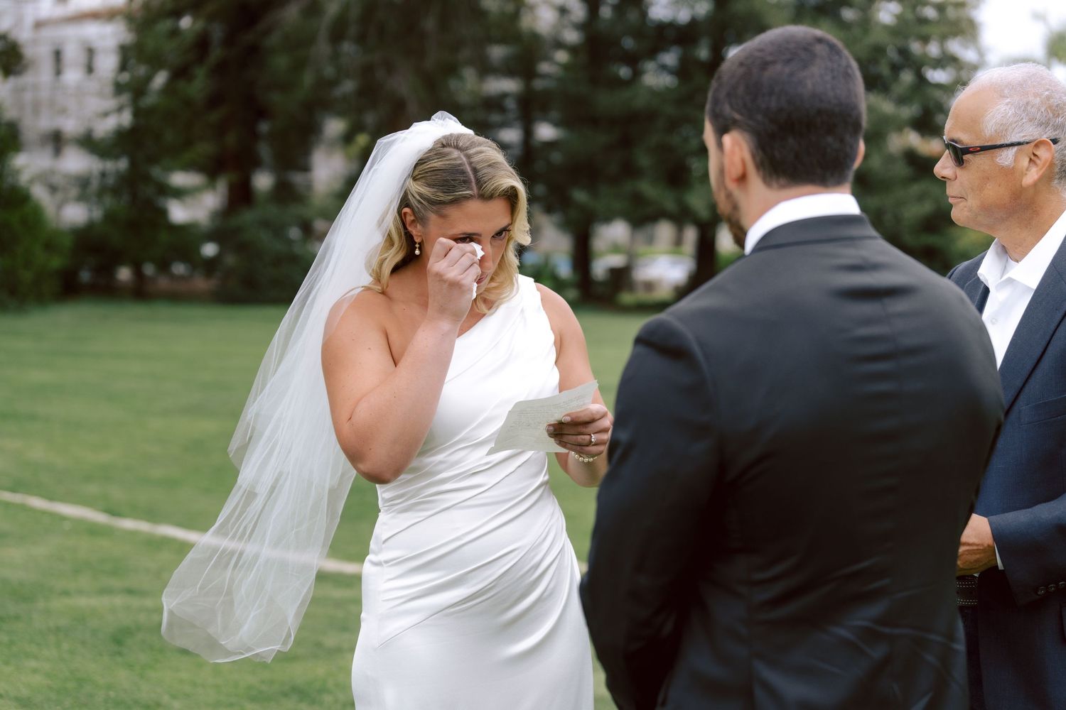 Emotional bride in white gown and veil wiping tears during outdoor wedding ceremony vow exchange.