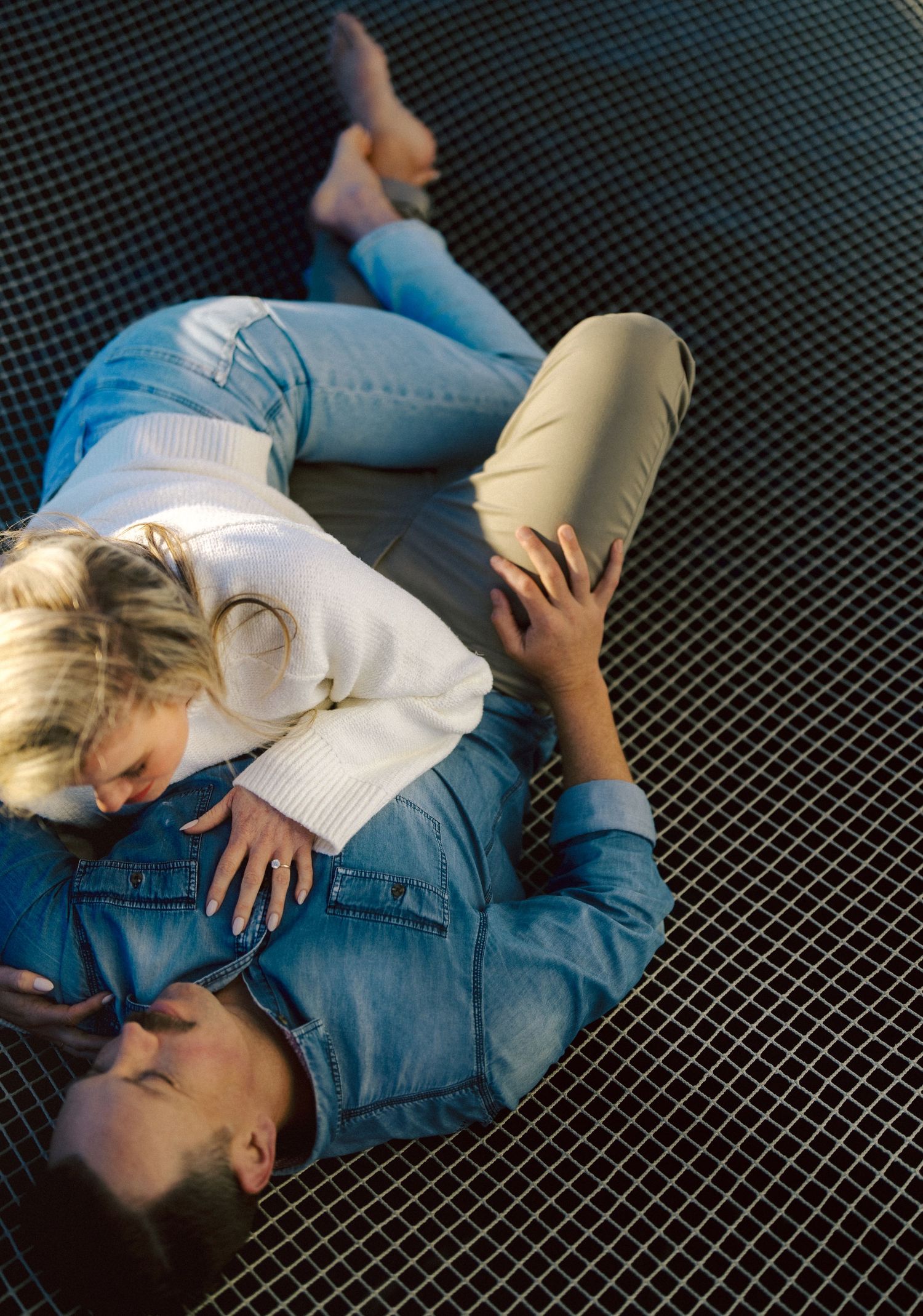 Couple lying together on a grid-patterned surface in soft sunlight during a relaxed engagement photo session.