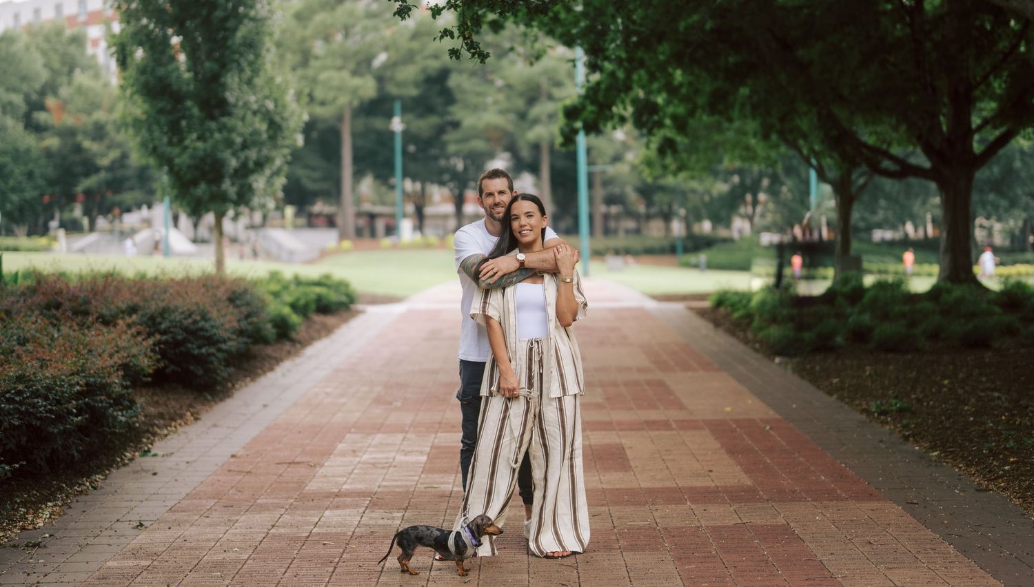 Smiling couple posing with their small dog on a brick park pathway surrounded by lush green trees.