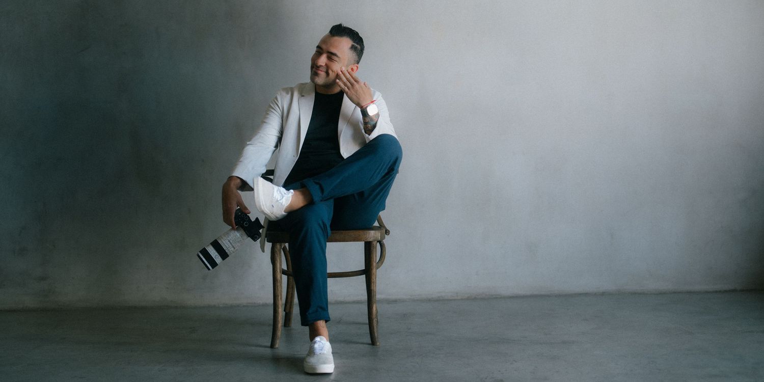 Photographer in white blazer sitting on chair holding camera against gray backdrop.