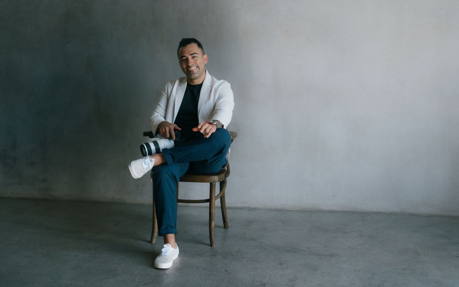 Man in white blazer and blue jeans sitting on wooden chair against gray concrete wall.