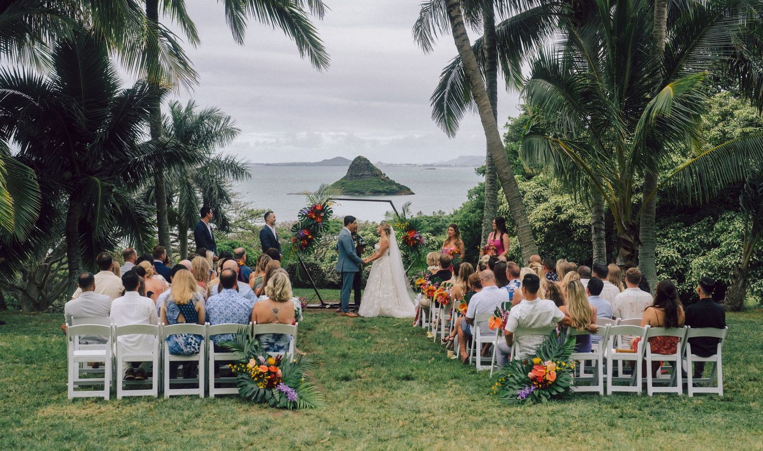 Outdoor tropical wedding ceremony with guests seated on white chairs, lush palm trees, and ocean view in background.