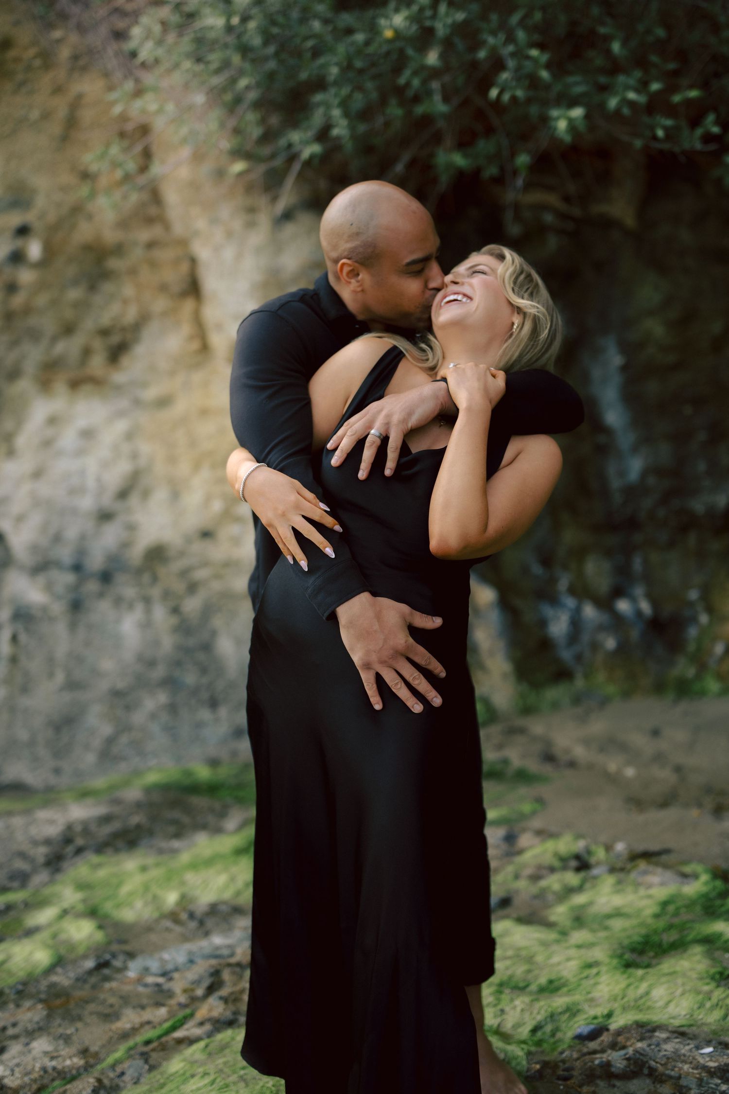 Loving interracial couple embracing and laughing together against mossy rock wall outdoors.